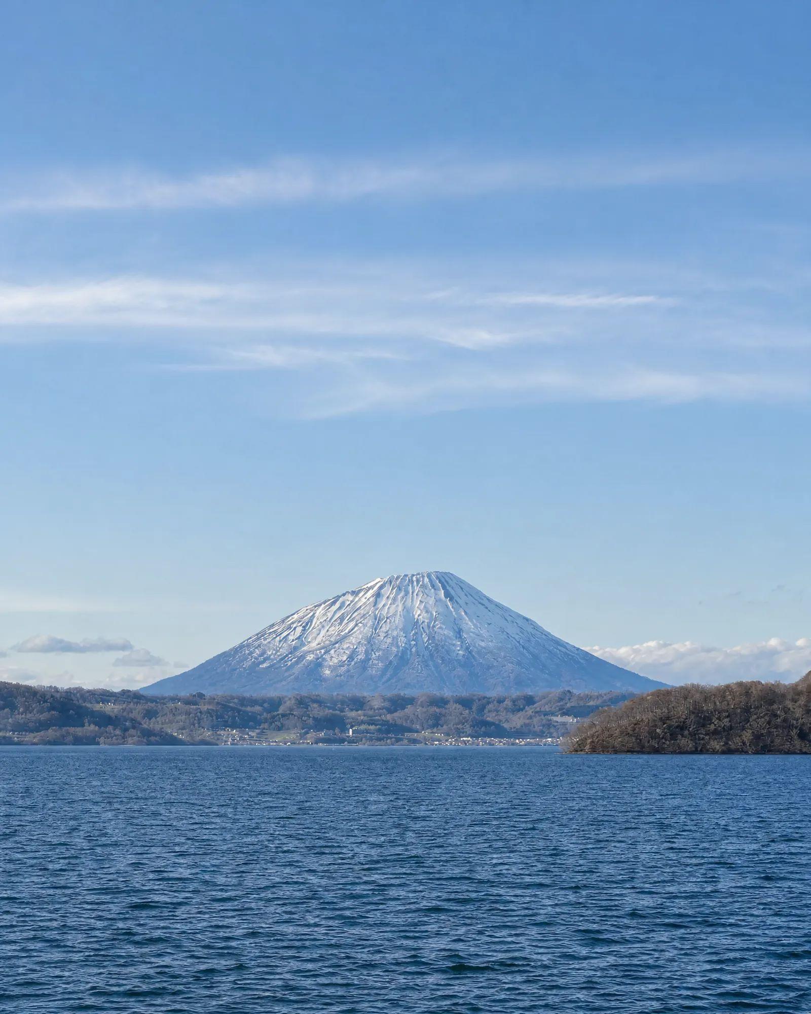 Snow-capped Mount Yotei rising beyond the blue waters of Lake Toya under a clear sky in Hokkaido, Japan.